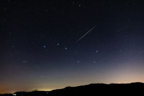 A night sky filled with stars and a shooting star streaking above silhouetted hills