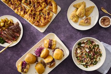 table with purple cloth and varerty of dishes, steak, sliders, grain salad, and more