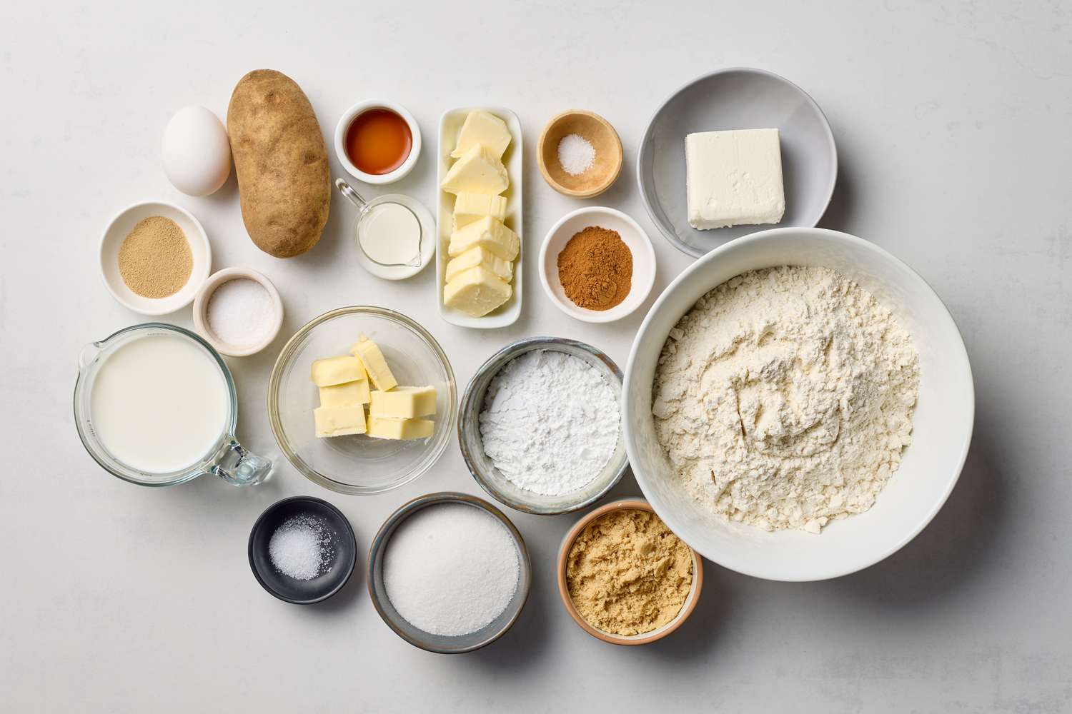 Ingredients for making cinnamon rolls arranged on a countertop including flour eggs butter milk sugar and spices like cinnamon