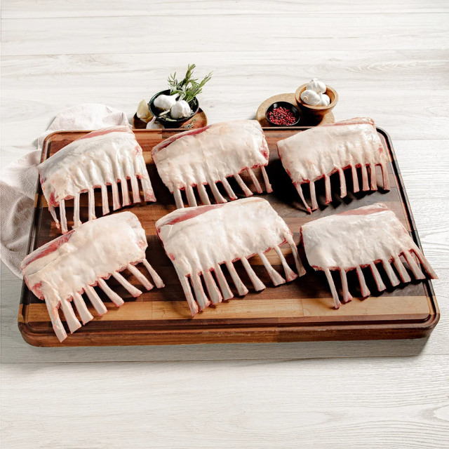 Rack of lamb portions arranged on a wooden board with seasoning and herbs in small bowls nearby