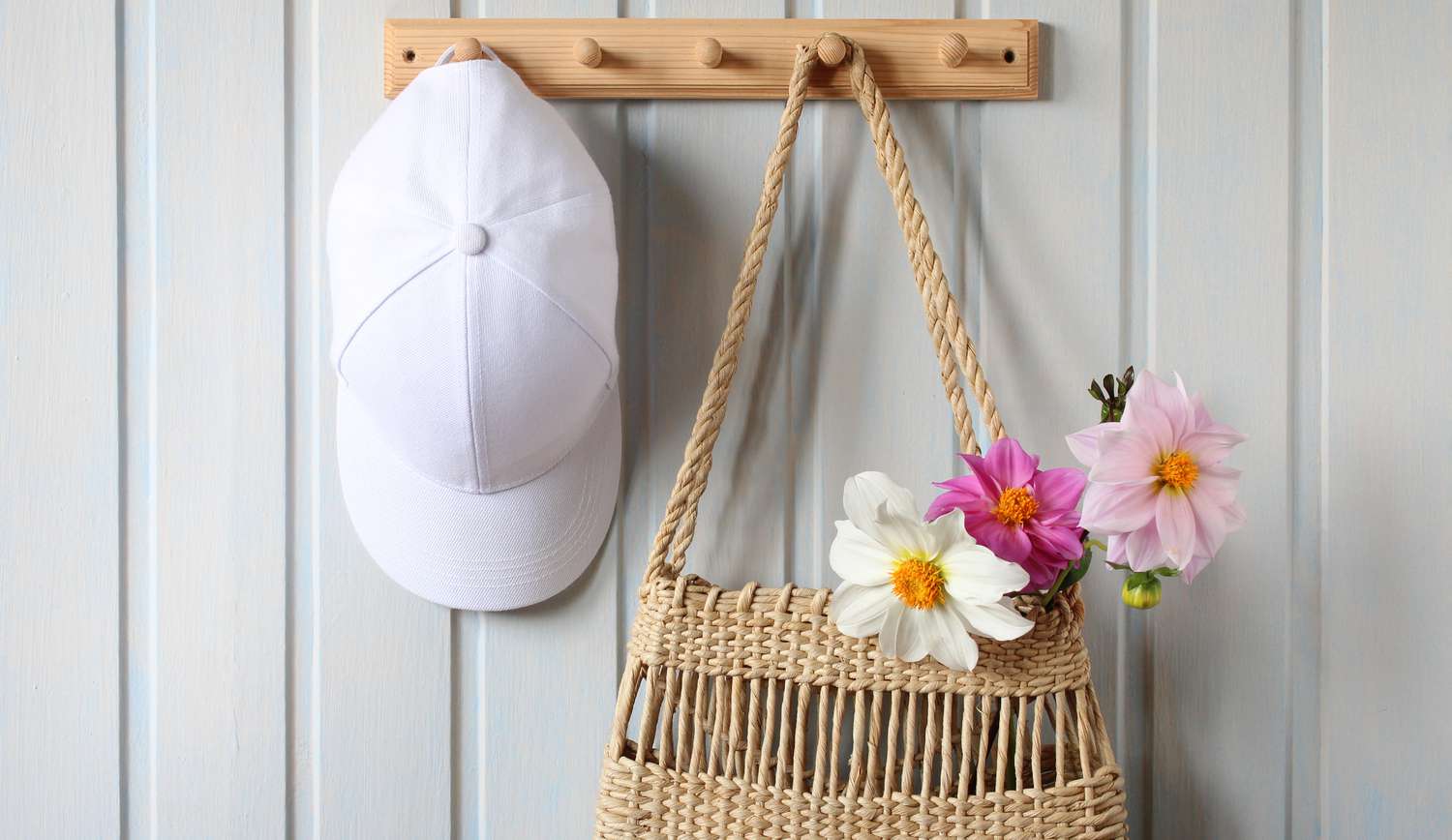 A wicker bag with a bouquet of dahlias and a white baseball cap hang on a wooden hanger on a board wall indoors.