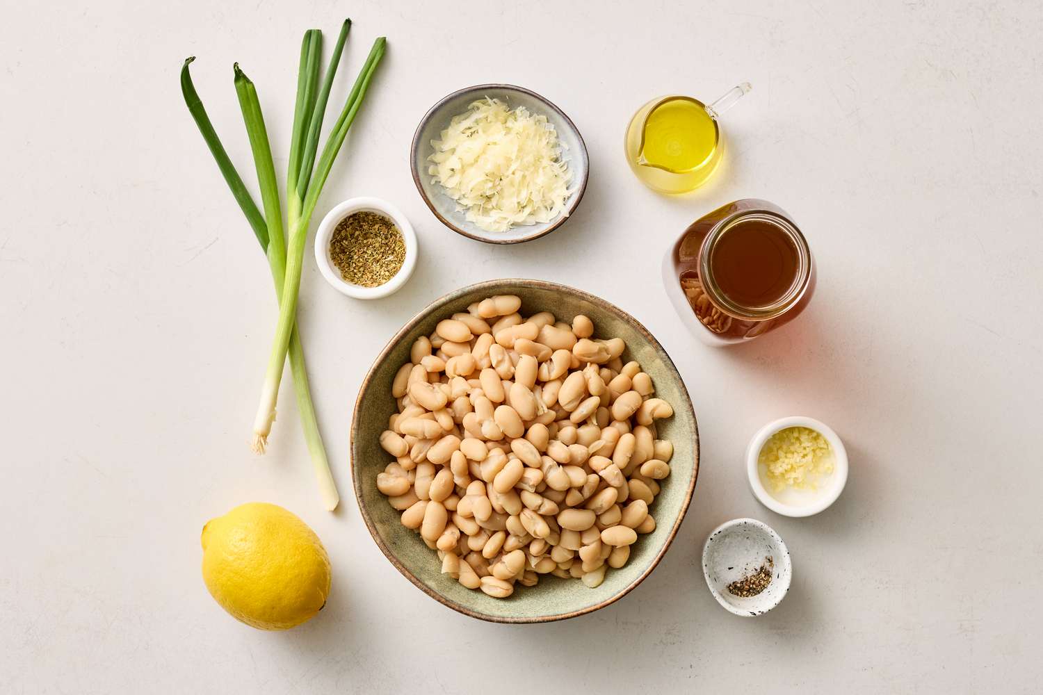 Ingredients for a white bean soup including a bowl of beans lemon green onions grated cheese olive oil broth and spices
