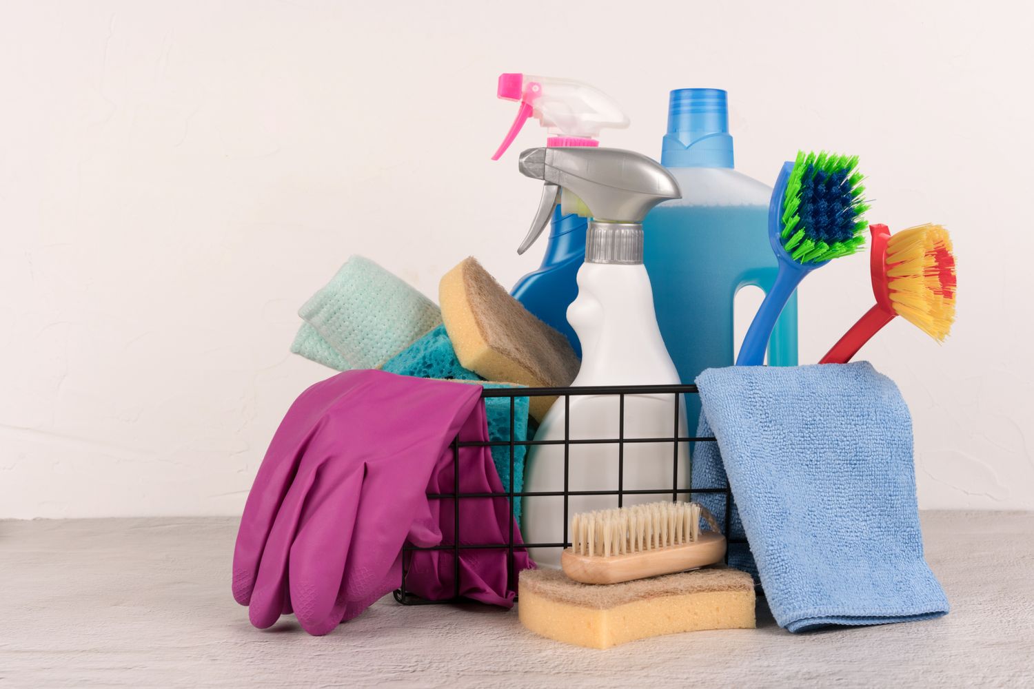 Various cleaning supplies including brushes spray bottle sponges towel and gloves in a storage basket