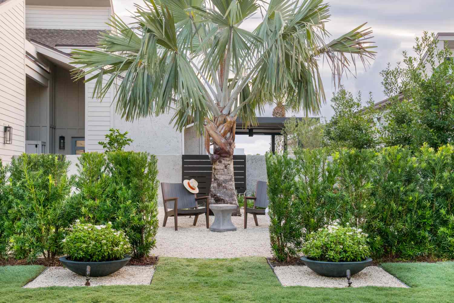 A landscaped garden featuring a palm tree a seating area with chairs and a table surrounded by trimmed hedges and greenery