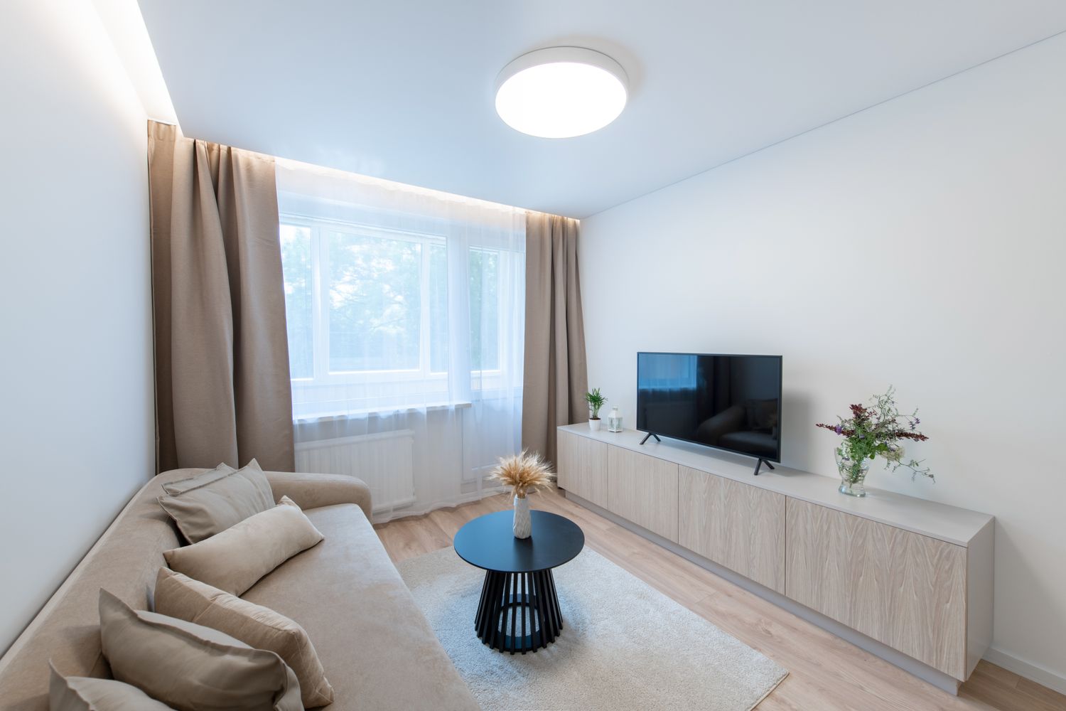 Modern Home Living Room Interior with Open TV Area and TV on the Wooden Desk, Sofa, And Coffee Table. Flowers on the Table. Windows and Curtains in Background. Hardwood Floor, Carpet. Wide Angle