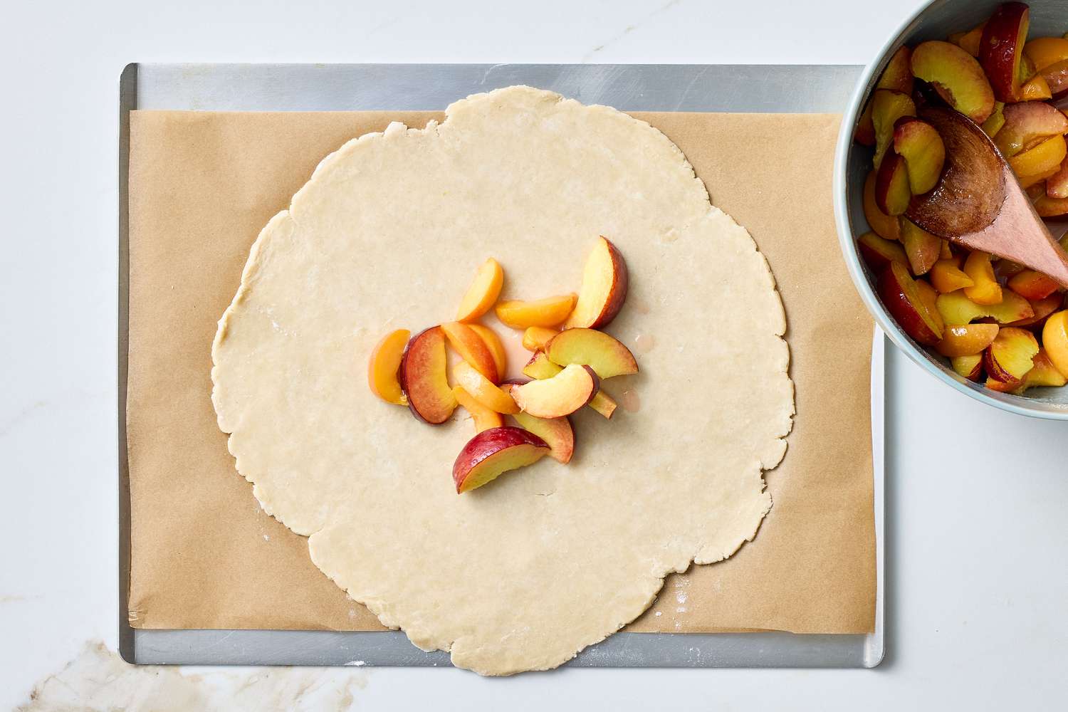 Rolled-out dough with sliced stone fruits on a baking sheet, a bowl with more fruit and spoon to the side