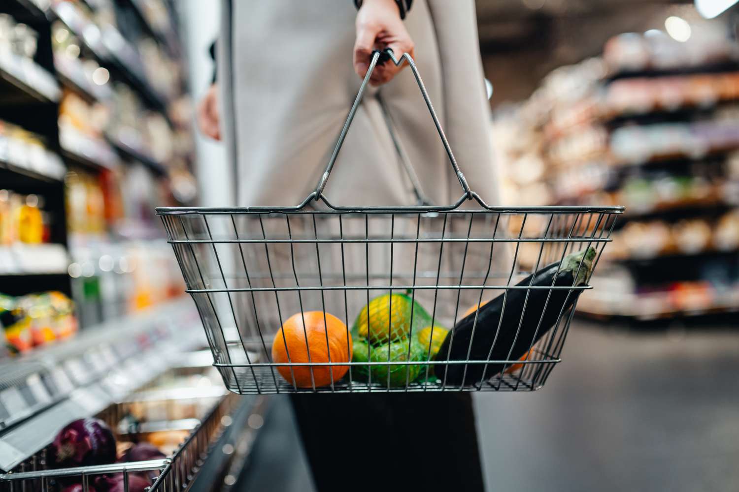 A person holding a shopping basket with fruits and a zucchini in a grocery store aisle