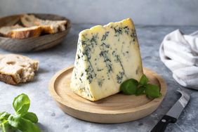A wedge of blue cheese with green leaves on a wooden platter bread in a bowl and a knife are nearby on the table
