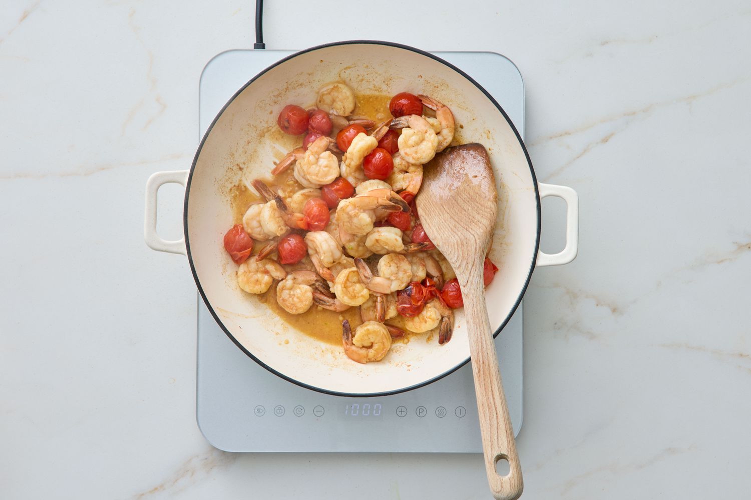 Sautéed shrimp with arugula and tomatoes in a pan on a stove, wooden spoon resting on the edge