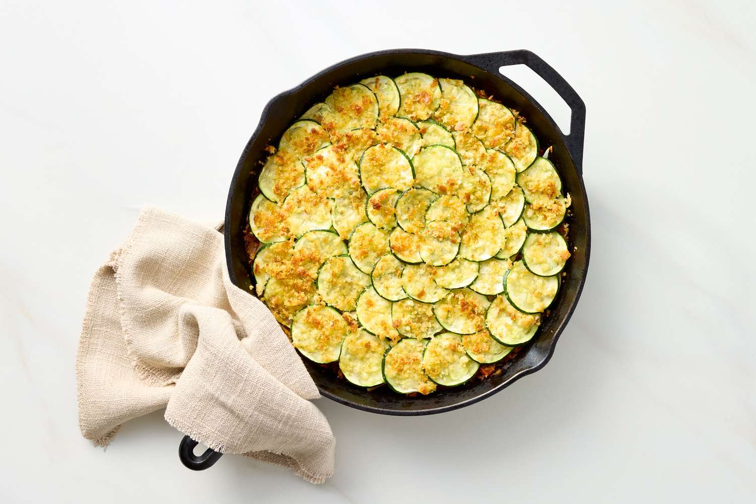A skillet containing a baked casserole with sliced zucchini and a cloth napkin on the handle