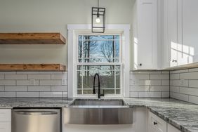 A modern kitchen sink area with a large window stainless steel sink and white cabinetry featuring open wooden shelves and a pendant light