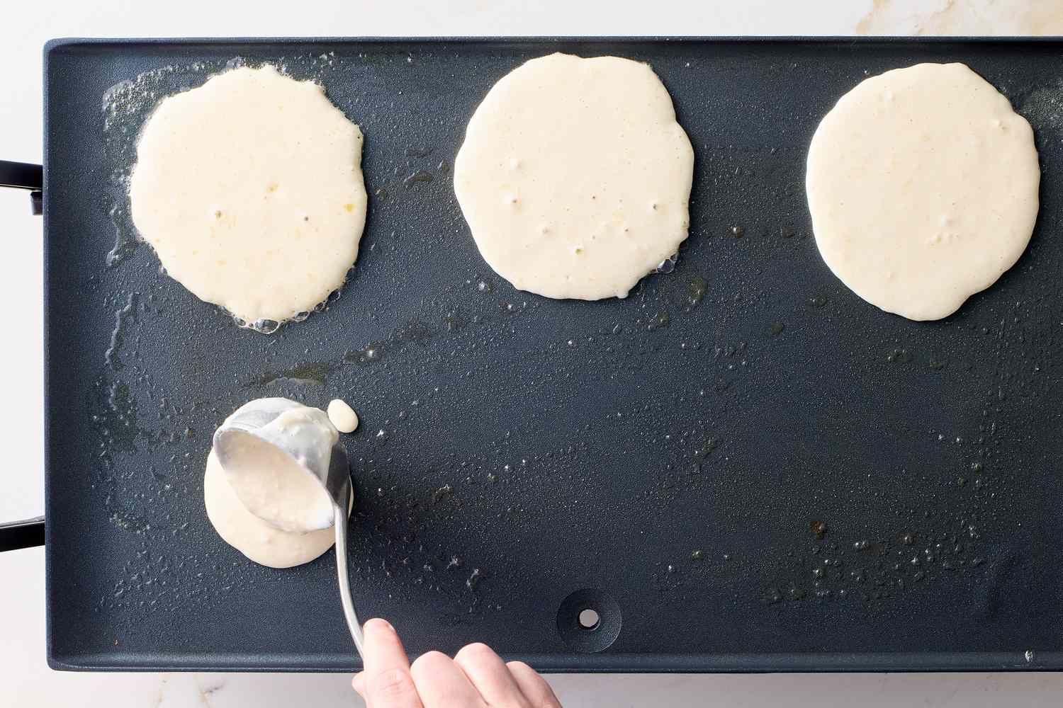 Pancake batter being poured onto a griddle with three pancakes cooking