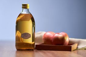 A bottle of apple cider next to a tray containing two apples on a wooden surface
