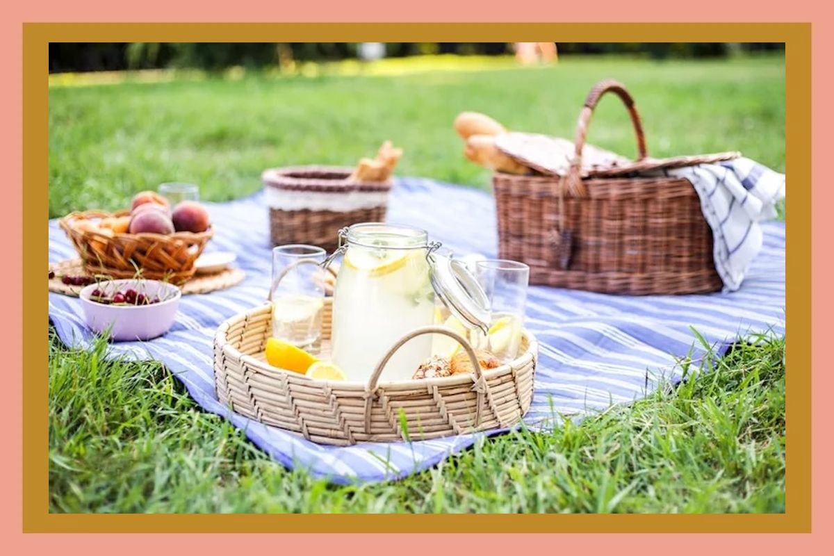 A picnic setup on a blanket featuring a tray with lemonade and glasses fruits in bowls and picnic baskets