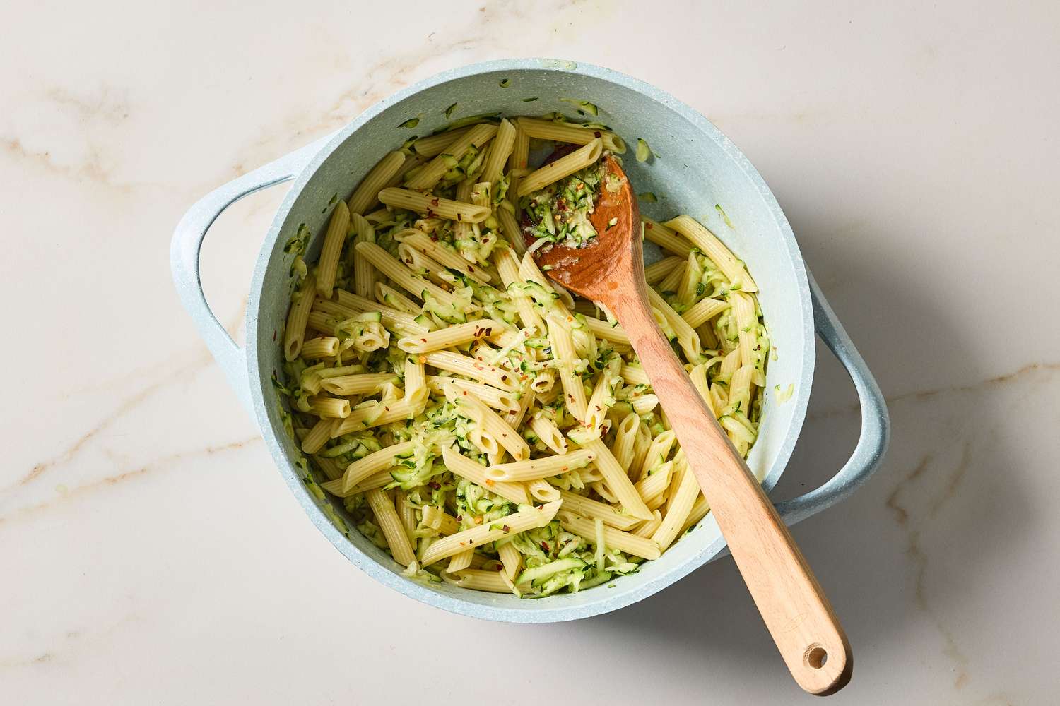 A pot containing cooked penne pasta mixed with shredded zucchini with a wooden spoon resting inside