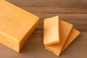 A block of cheddar cheese with three slices placed beside it on a wooden surface