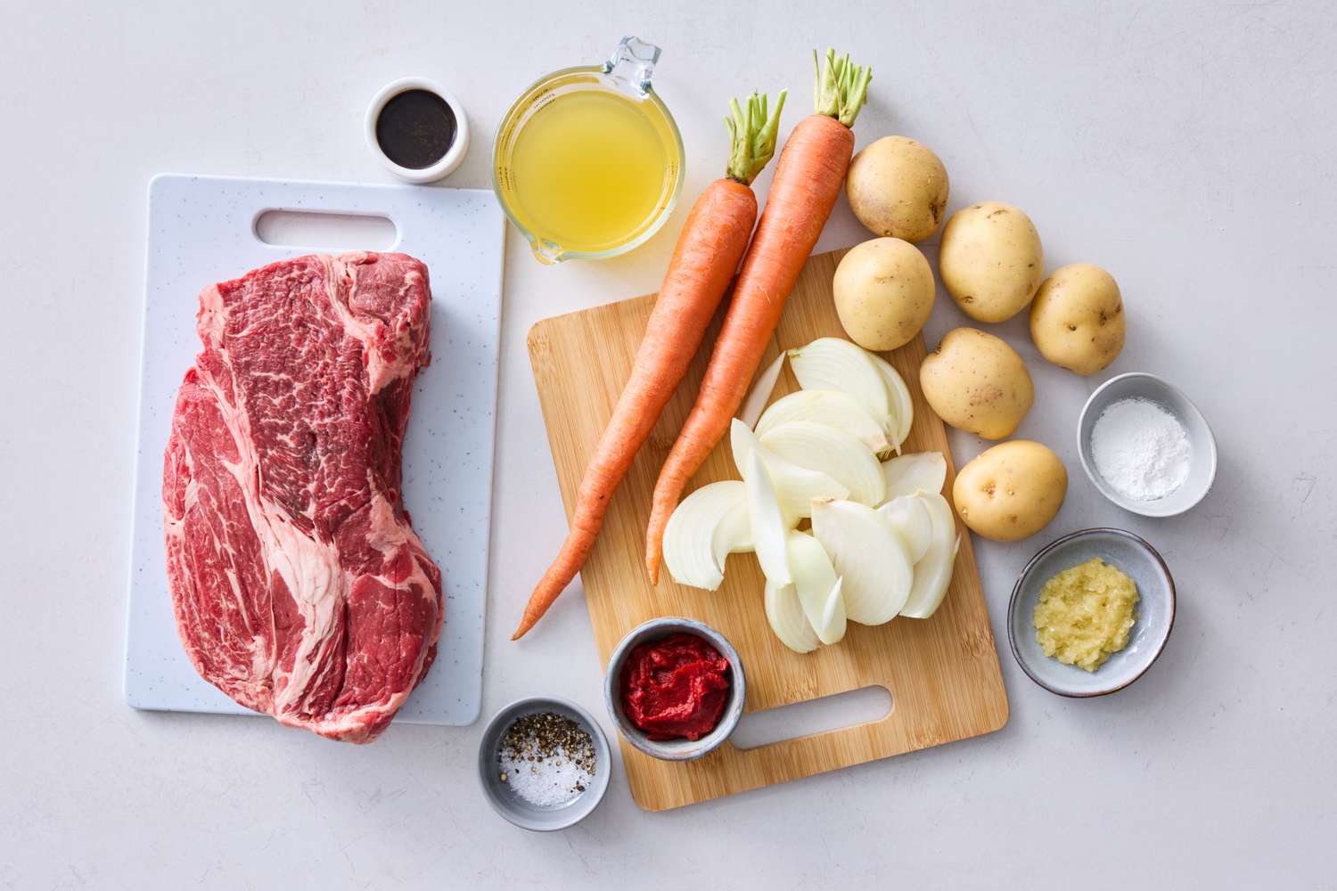 Ingredients for a pot roast including raw beef carrots potatoes onion and various small bowls with seasonings and liquids