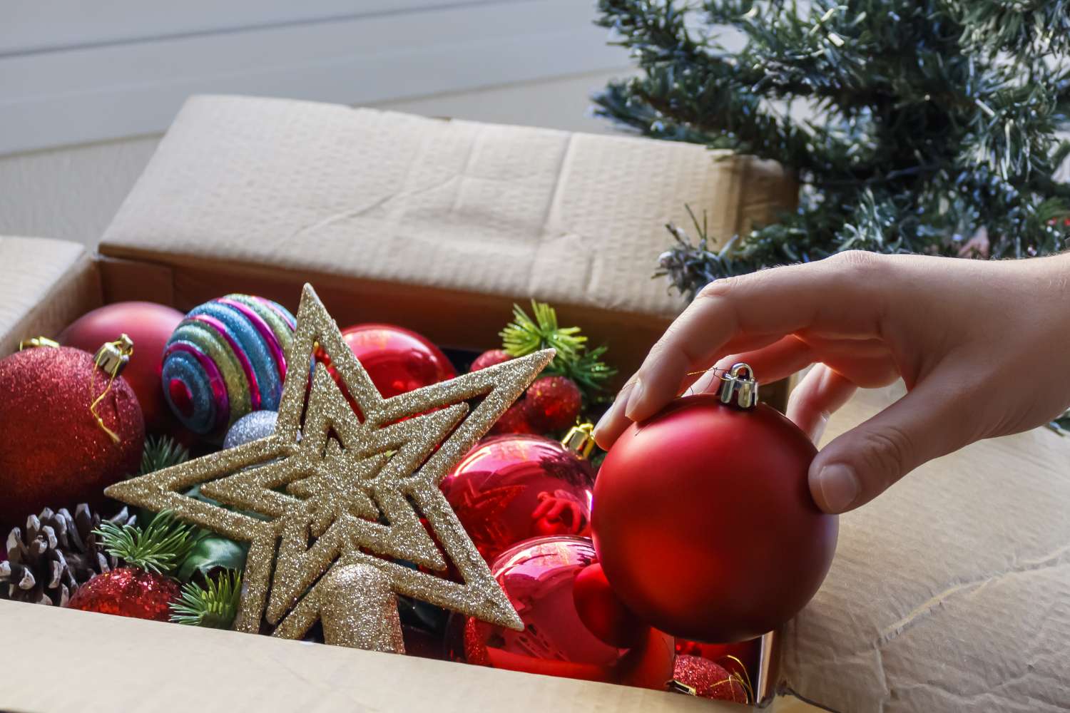 A hand reaching for a red bauble among various holiday ornaments in a box with a partially visible Christmas tree nearby