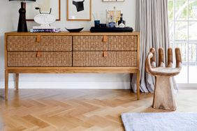 A wooden sideboard with woven drawers and a decorative wooden chair shaped like a hand in a room with a window