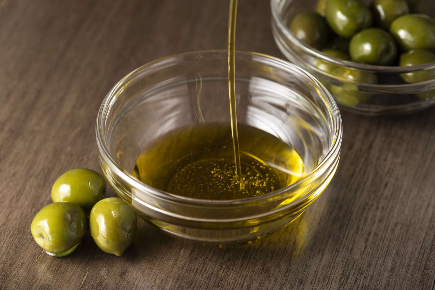 A closeup of olive oil being poured into a small glass bowl with fresh green olives nearby on a wooden surface