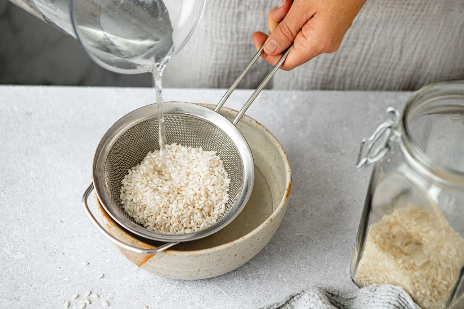 Hand rinsing uncooked rice in a metal sieve with a jar of rice on the side