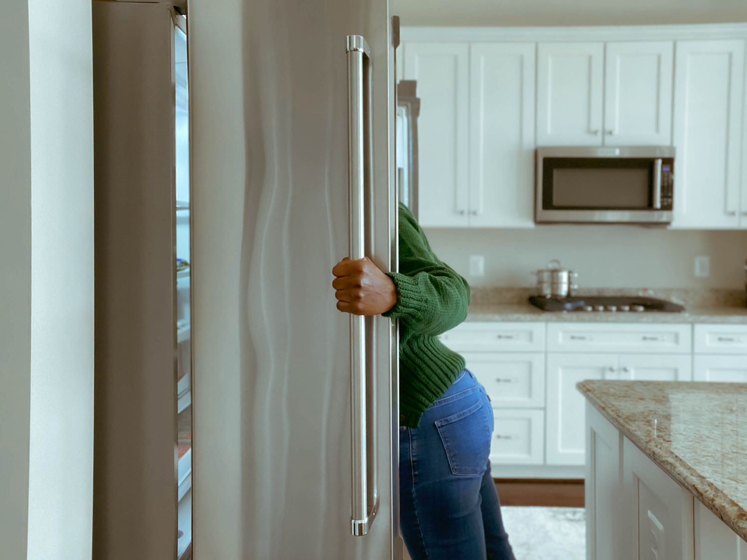 A person standing partially in view while opening the door of a refrigerator in a kitchen with white cabinetry
