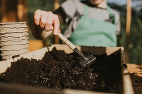 A person working with soil in a gardening environment using a tool above a tray filled with soil