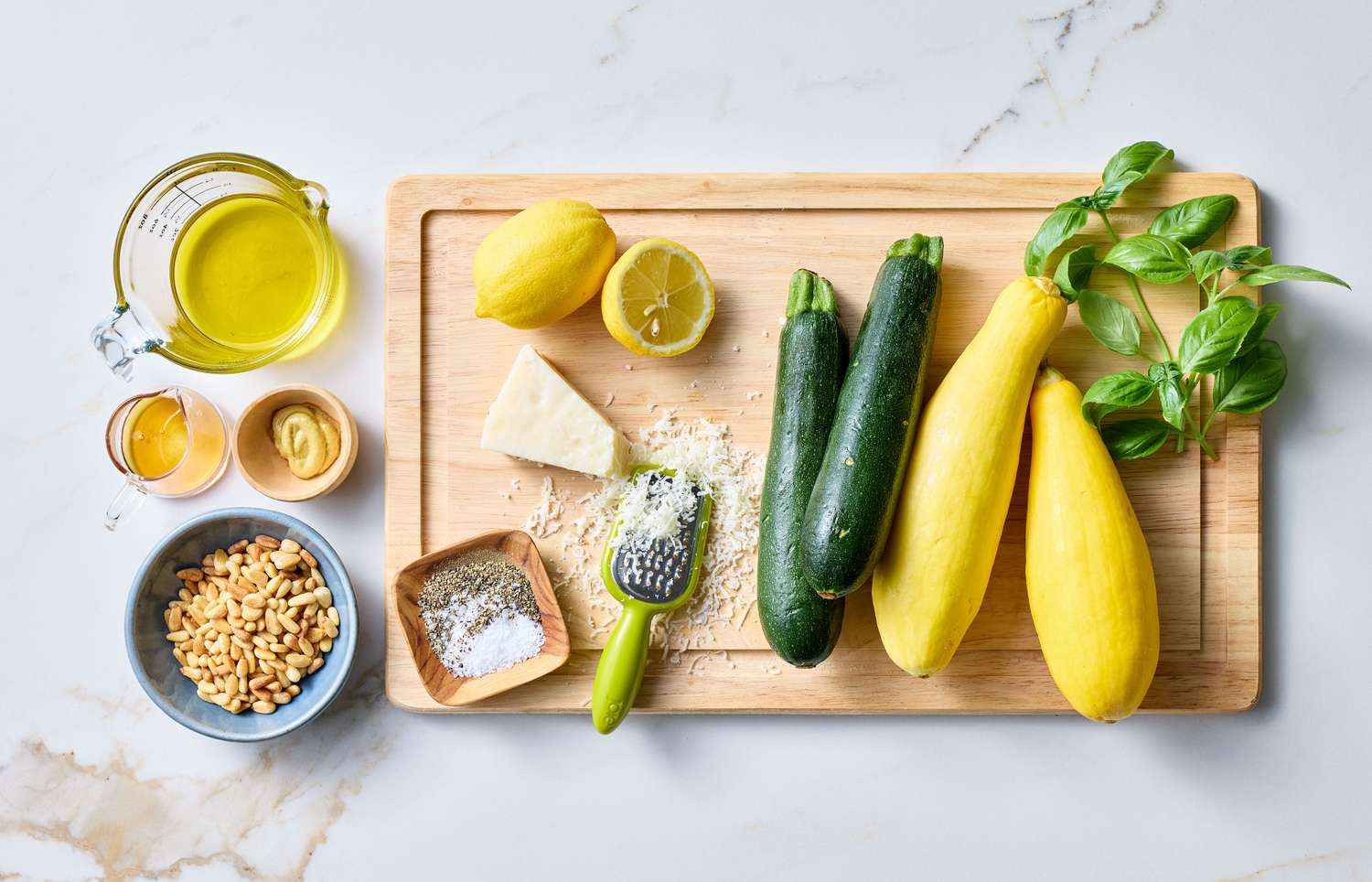 Ingredients for zucchini squash salad including zucchini, yellow squash, Parmesan cheese, olive oil, pine nuts, mustard, and basil on a cutting board