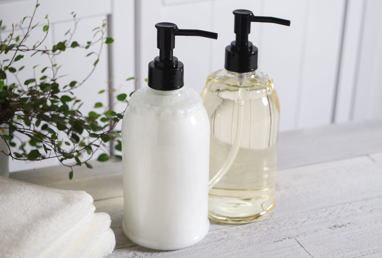 Two soap dispensers on a bathroom counter one filled with white soap and the other with a clear liquid near folded towels and a plant