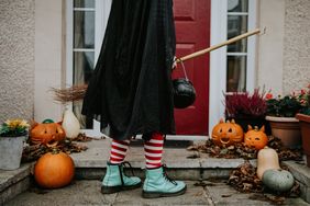 Person in witch costume with green boots holding a broom and cauldron standing in front of a decorated doorway with pumpkins and jackolanterns