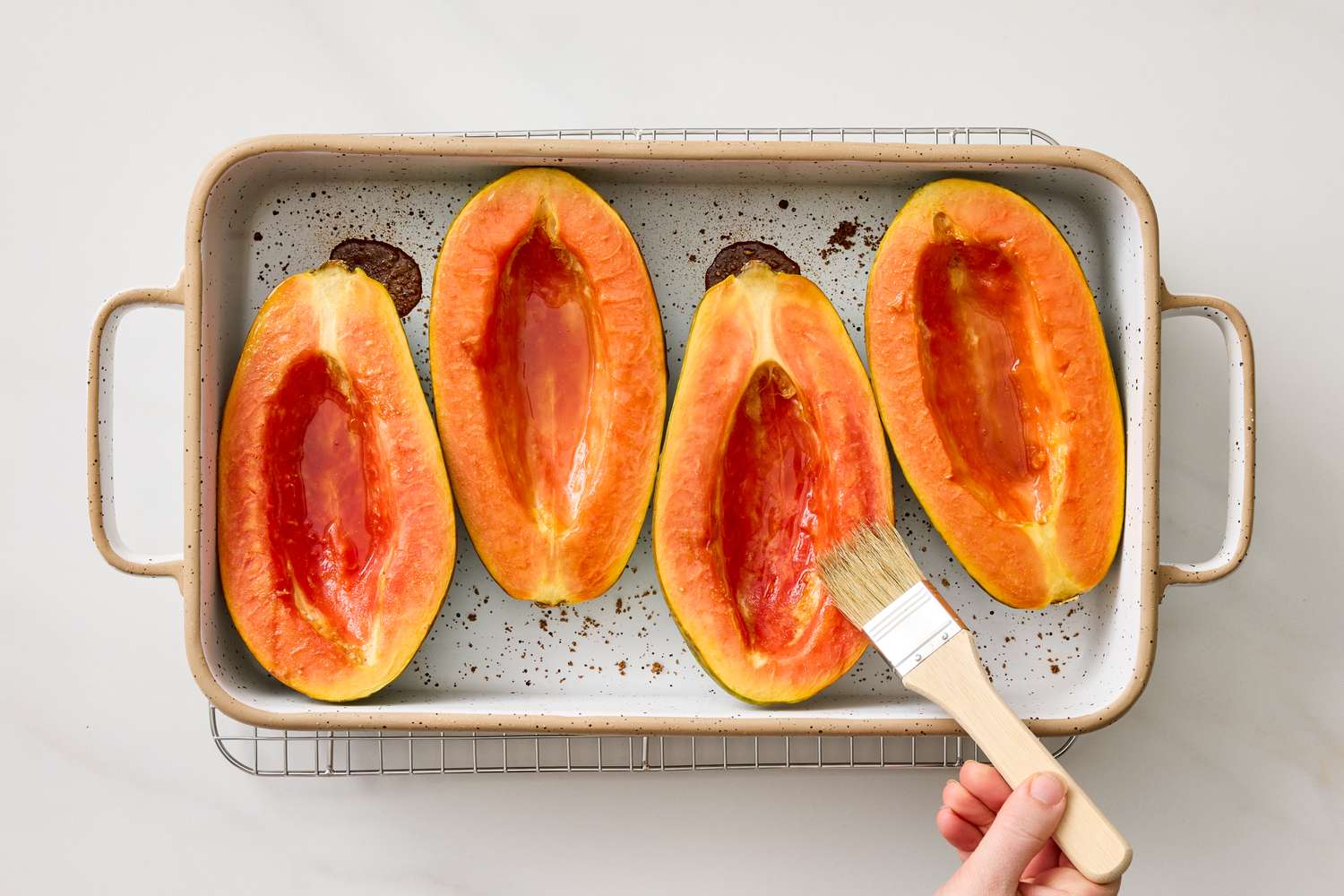 Four halved papayas placed on a baking tray being brushed with a liquid by a hand holding a pastry brush