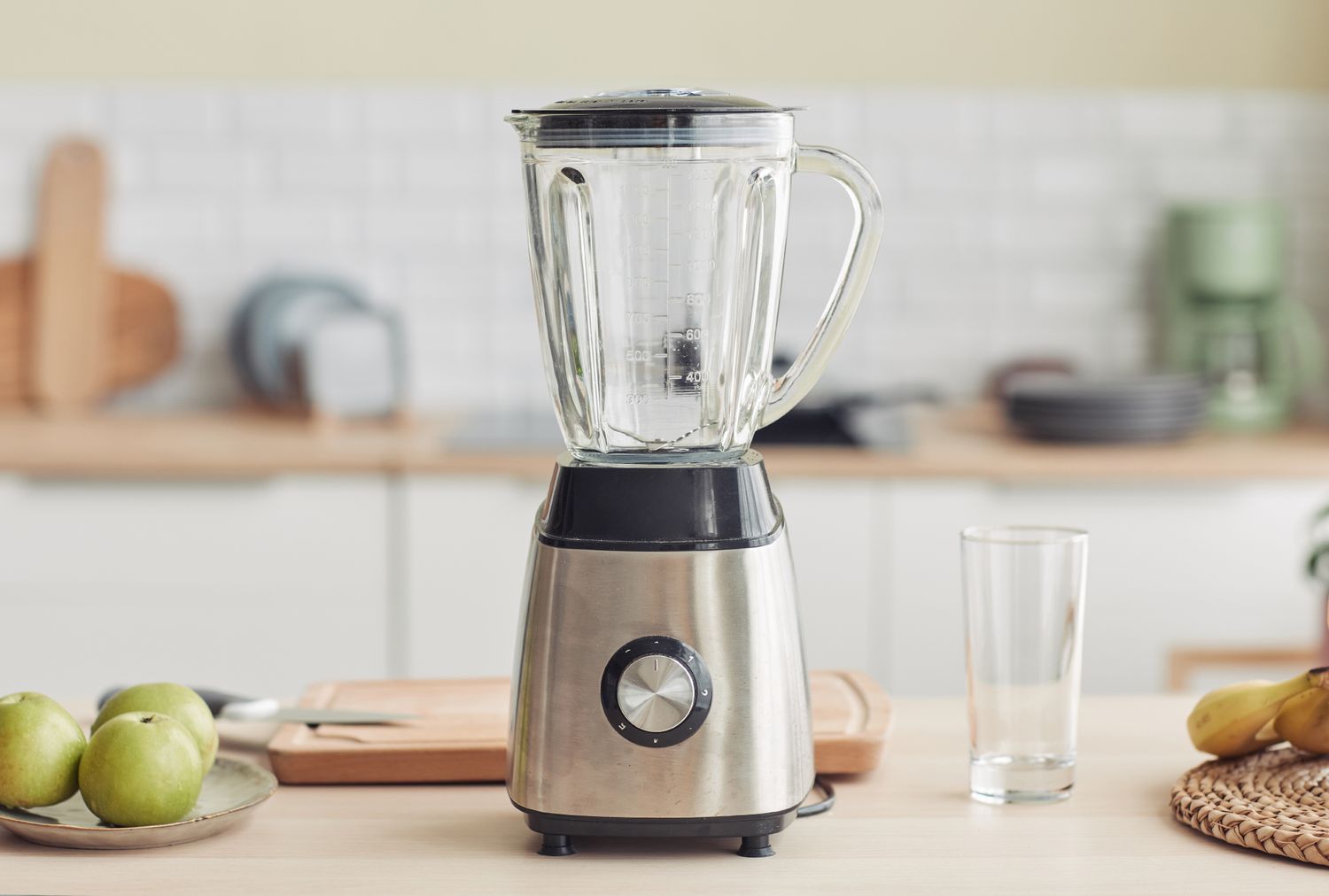 A kitchen blender placed on a counter in a home kitchen setting with various items like green apples and a drinking glass nearby