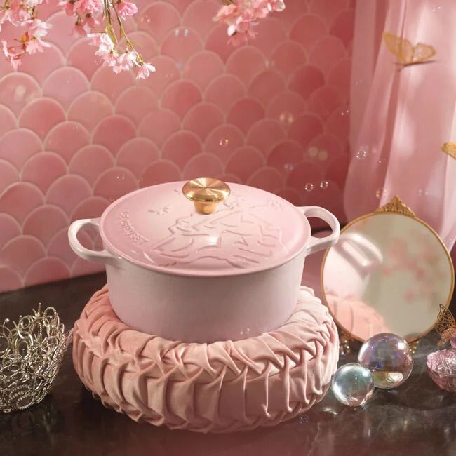 A pink cookware pot with a lid placed on a decorative cushion surrounded by reflective glass items and a mirror with a delicate backdrop