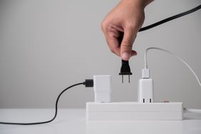 A hand plugging a power cord into a power strip next to several already connected devices