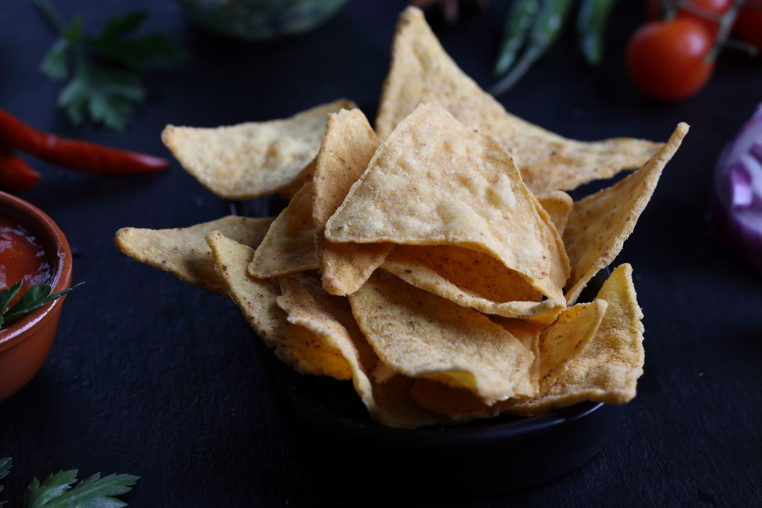 A bowl of tortilla chips with garnishing and vegetables on a dark surface