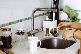 A person rinsing a cup under a kitchen faucet beside a sink with a mug and jar on the countertop