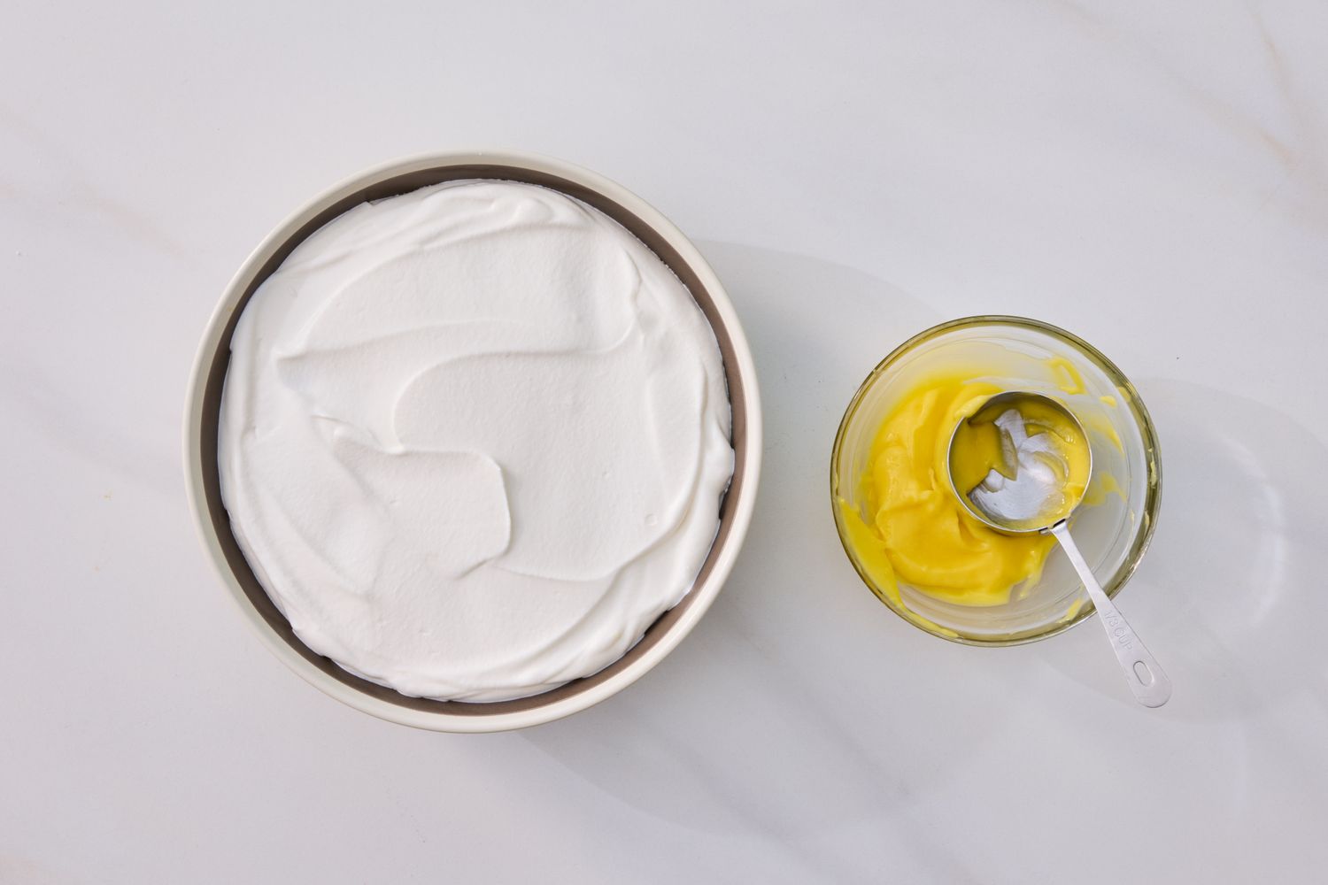 A bowl of whipped cream and a glass bowl with lemon curd and a spoon placed on a flat surface