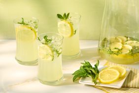 Three glasses filled with lemonade garnished with slices of lemon and mint with a pitcher and a plate of lemon slices nearby