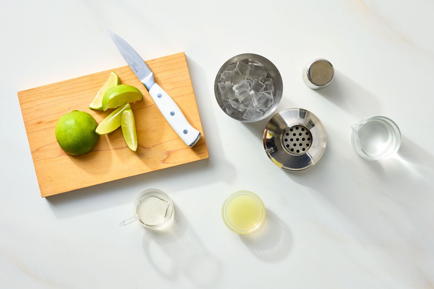 Cocktailmaking setup including lime wedges a cutting board knife metal strainer ice and small containers of liquids