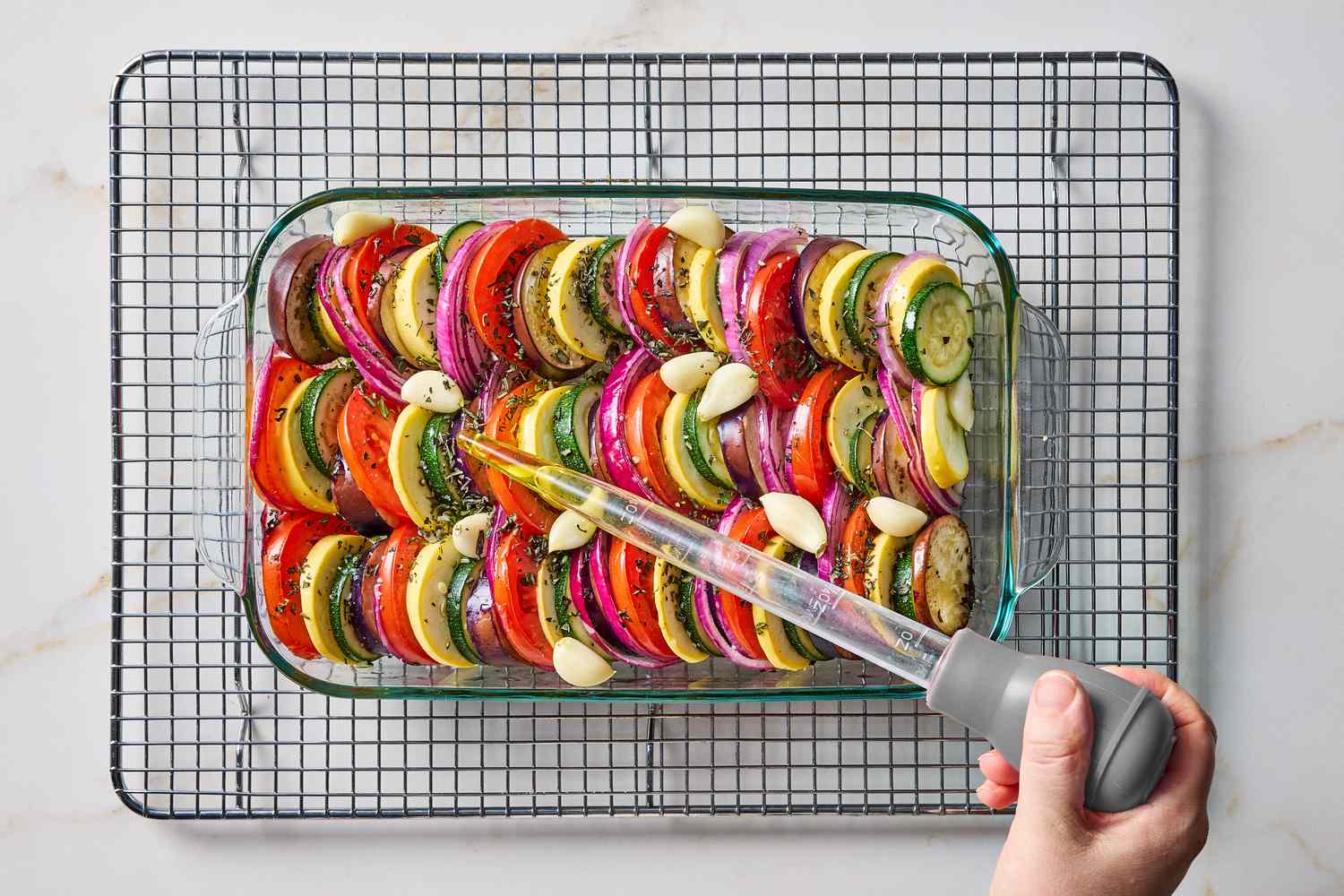 A hand drizzling oil with a baster over sliced vegetables in a baking dish