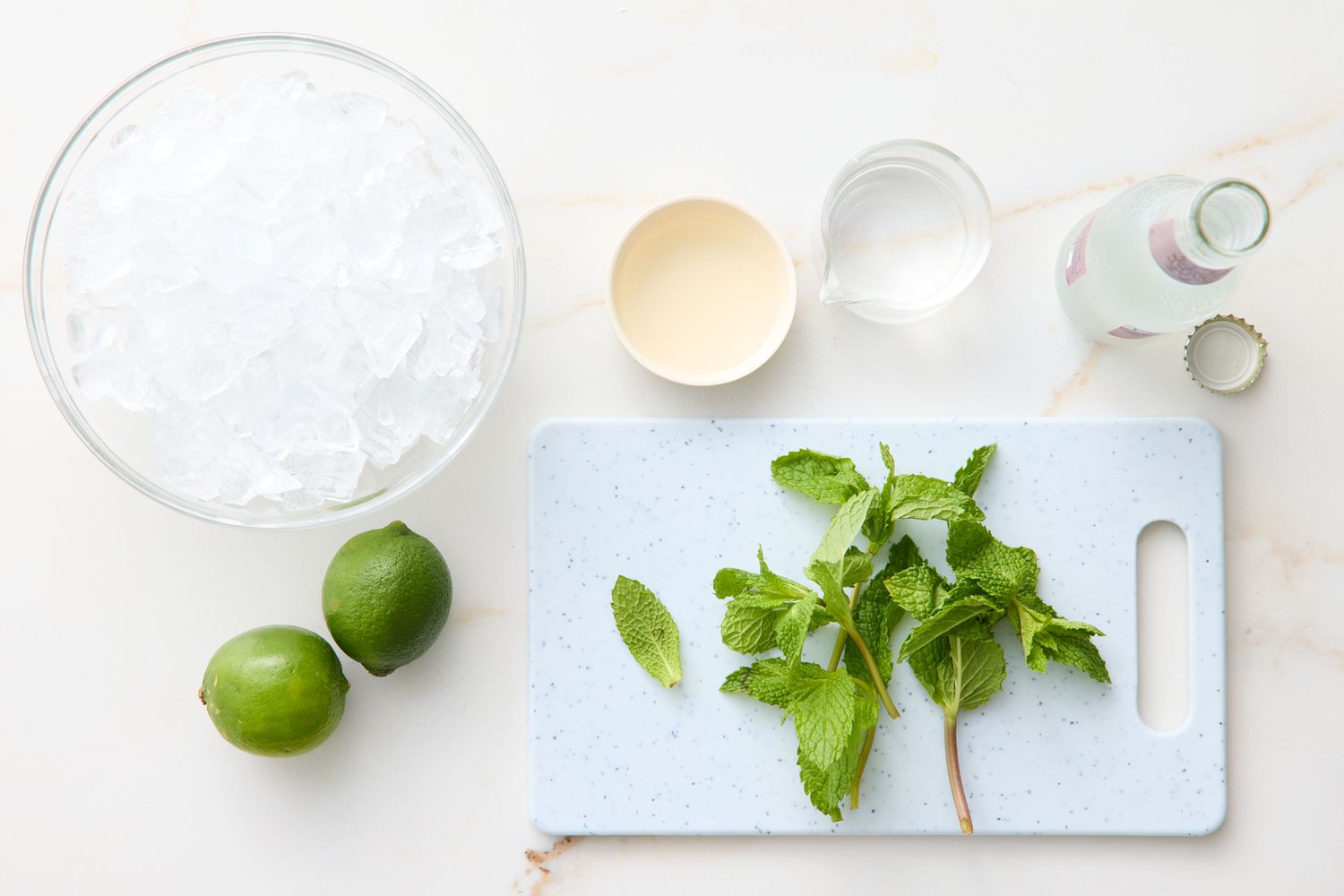 Ingredients for a mojito, including limes, mint leaves, a bowl of ice, liquid ingredients, and a bottle of carbonated water on a countertop