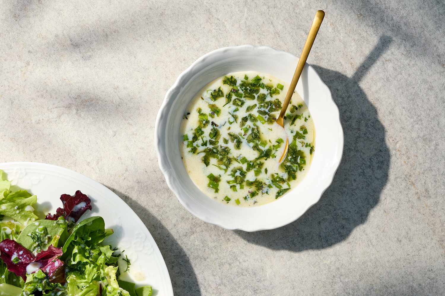 A bowl of buttermilk herb vinaigrette with a golden spoon and a portion of salad on a white plate