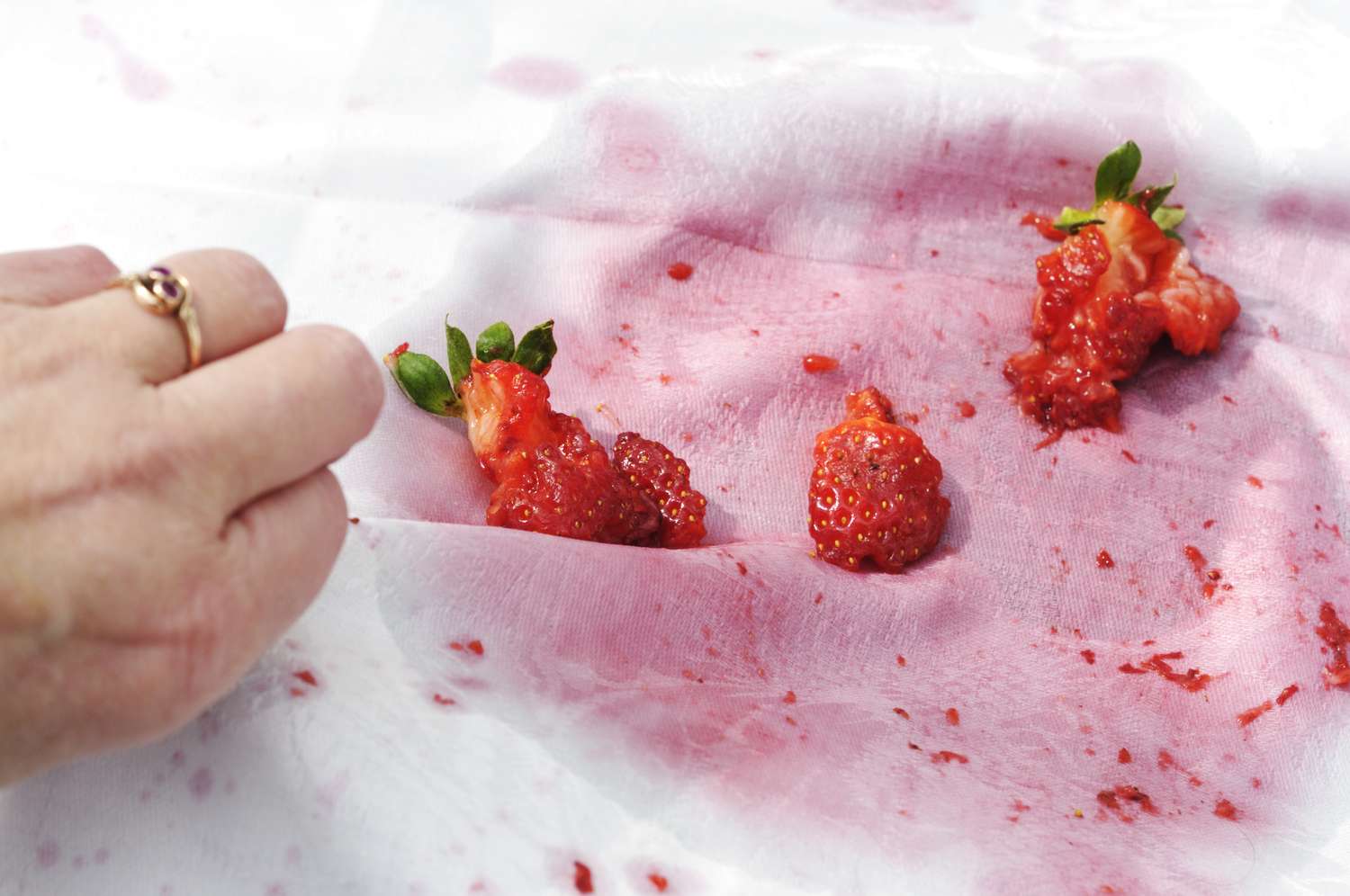 A hand holding fabric stained with mashed strawberries and juice placed on a white surface