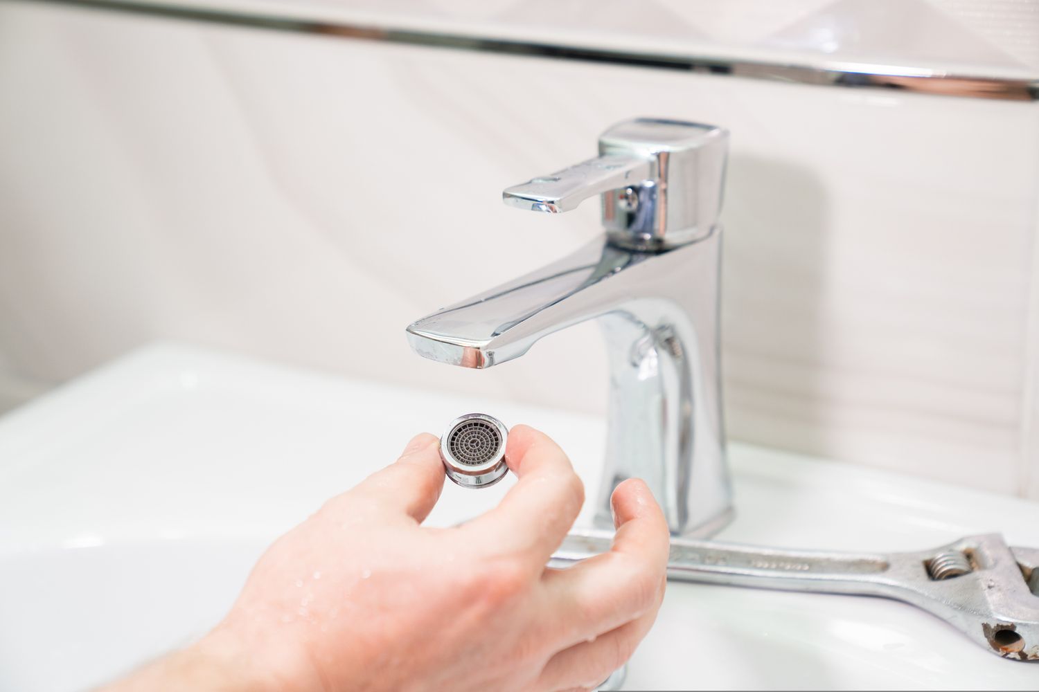 Hand holding a faucet aerator next to a sink with a wrench visible