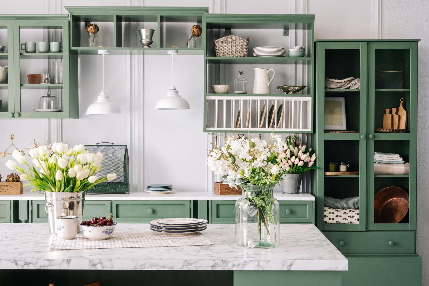 Kitchen with green vintage furniture, marble countertop with flowers and bowl of cherries, bucket with tulips, cupboard with various mugs, crockery and devices.