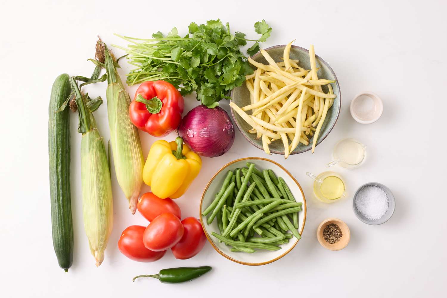 Various fresh salad ingredients including peppers tomatoes onions corn green beans and herbs arranged on a white surface