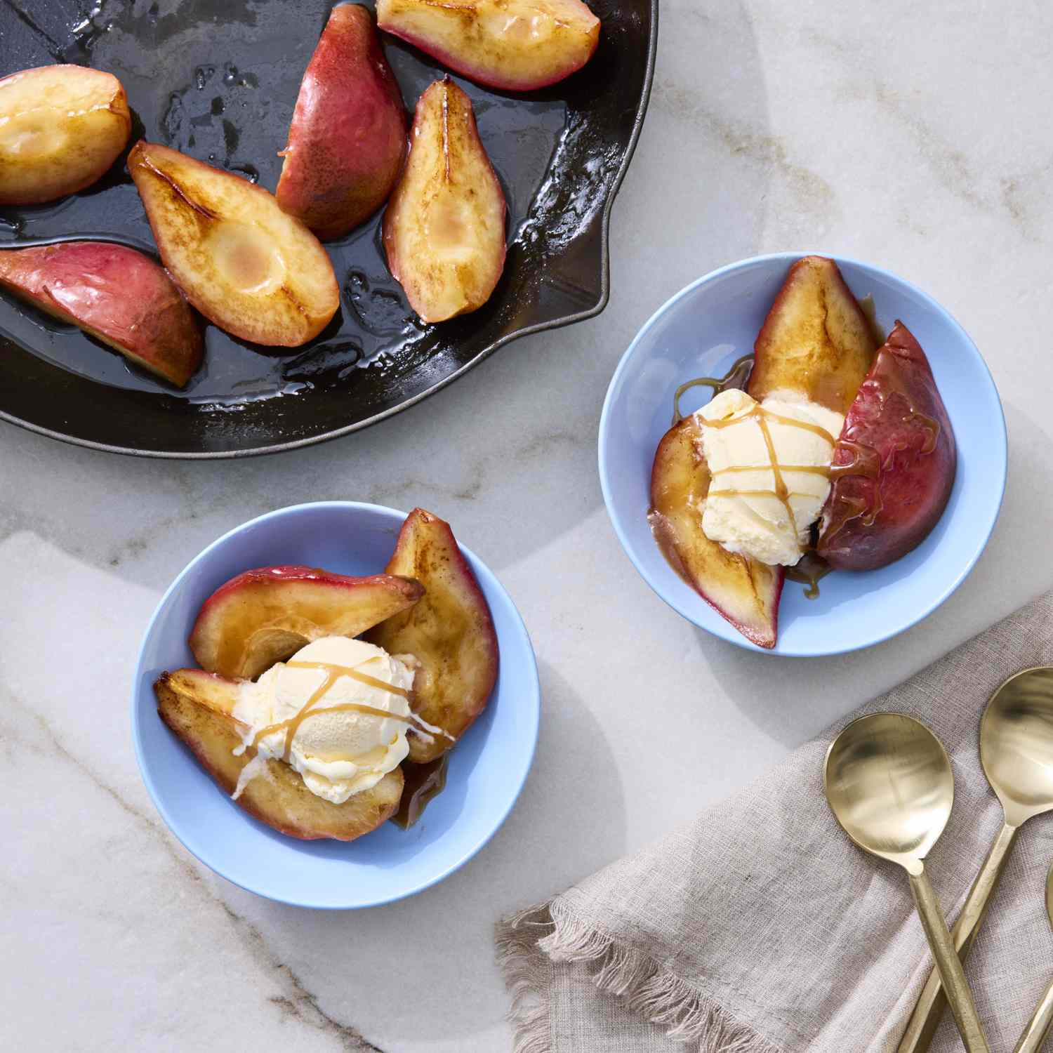 Caramelized pear halves with served scoops of ice cream and caramel drizzle plates on a table with a skillet and spoons nearby