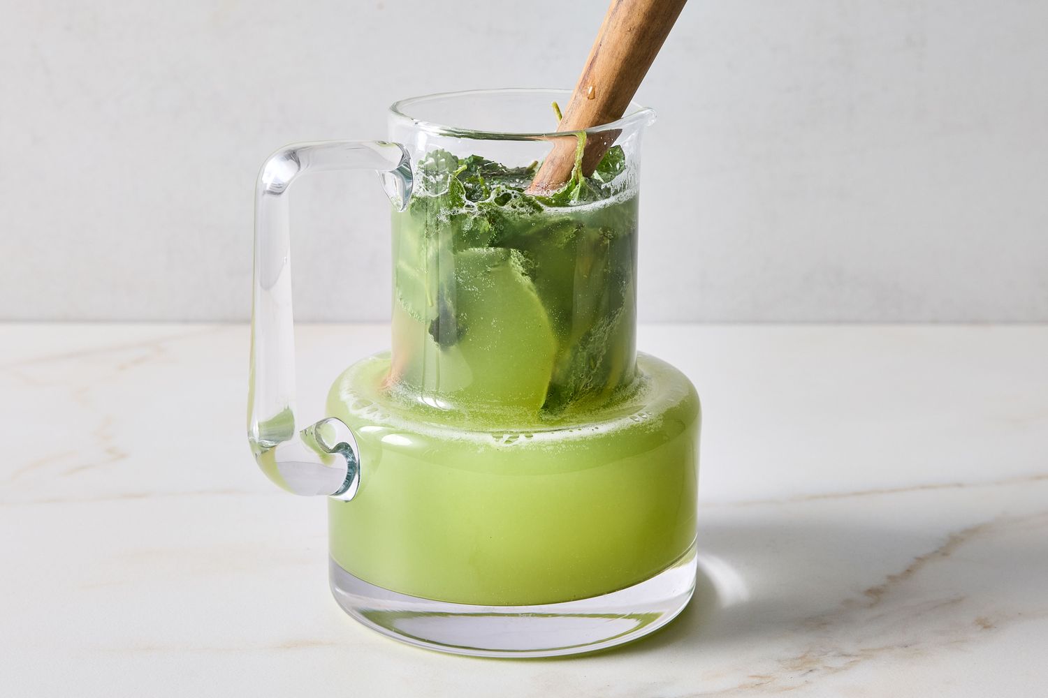 Glass pitcher containing cucumber cooler beverage with herbs and a wooden muddler