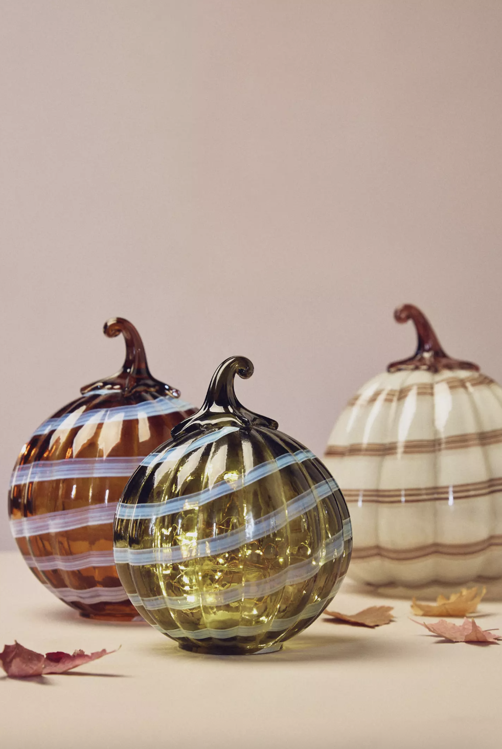 Glass pumpkins with stripe patterns displayed on a surface surrounded by scattered autumn leaves