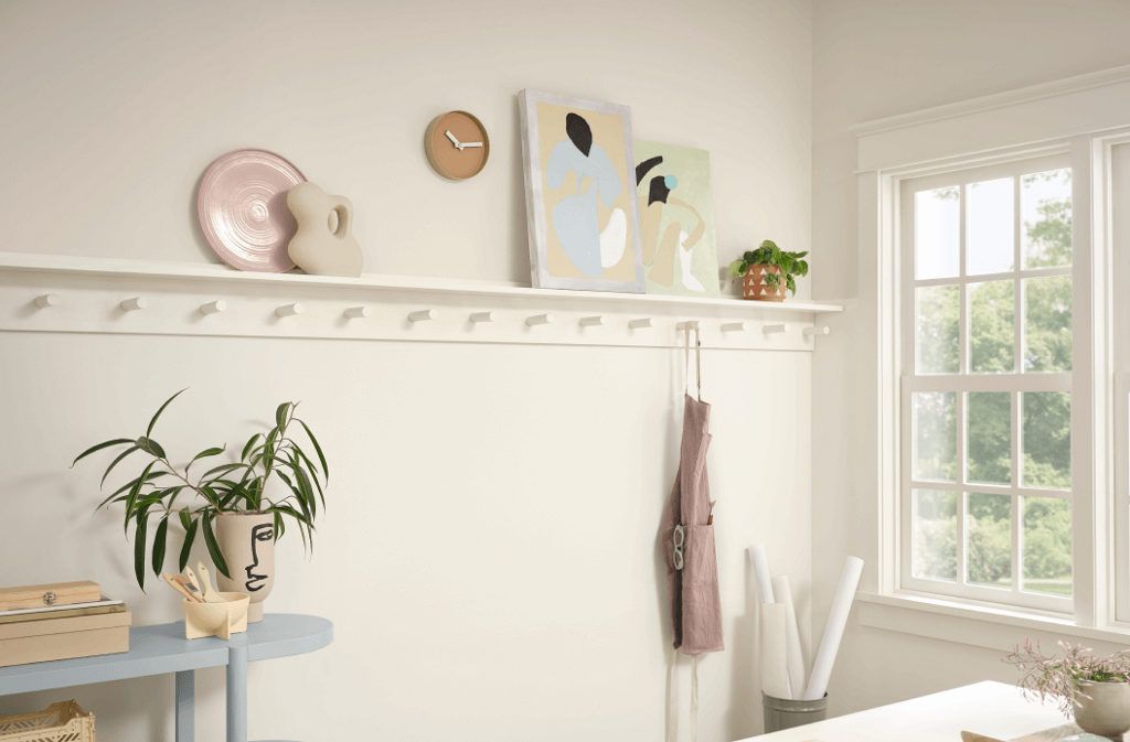 A home workspace featuring a shelf with artwork pottery plants and household items along with hanging apron near a sunlit window